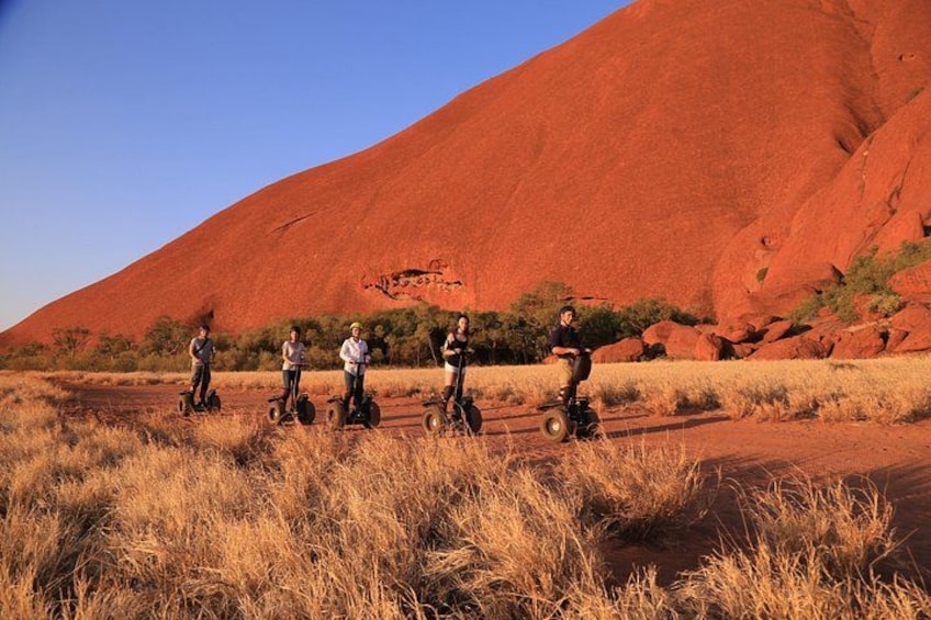 A group of people riding Segways in a dry grassland with a large red rock formation in the background. 4