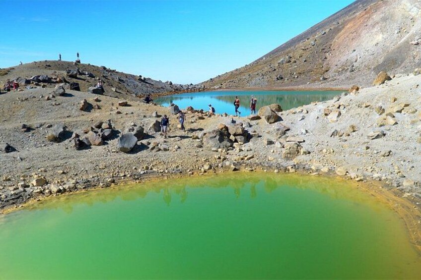 A rocky landscape with a greenish pool of water, a clear sky, and a few people standing on the shore. 4
