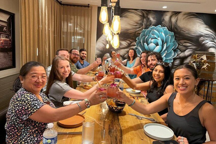 Un grupo de personas brindando con bebidas alrededor de una mesa de madera en un restaurante con un mural floral de fondo. 2