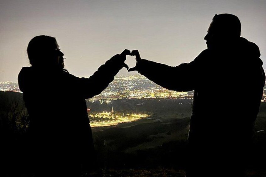 Dos personas formando una forma de corazón con las manos frente a un paisaje urbano al anochecer. 3