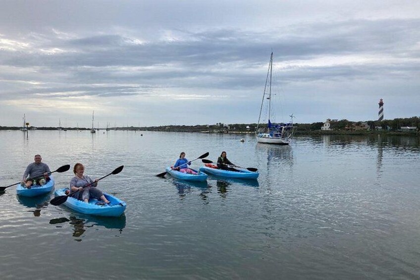 People kayaking on calm water with a sailboat and a lighthouse in the background. 2