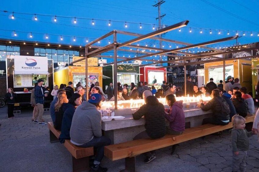 Un marché alimentaire où l’on peut voir des gens assis à des tables, des étals de nourriture et une tente illuminée de guirlandes lumineuses. 3