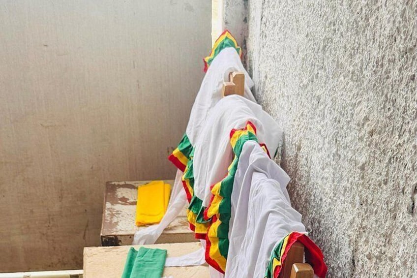 Una persona vestida con una prenda blanca y adornos coloridos apoyada contra una pared con textura. 3