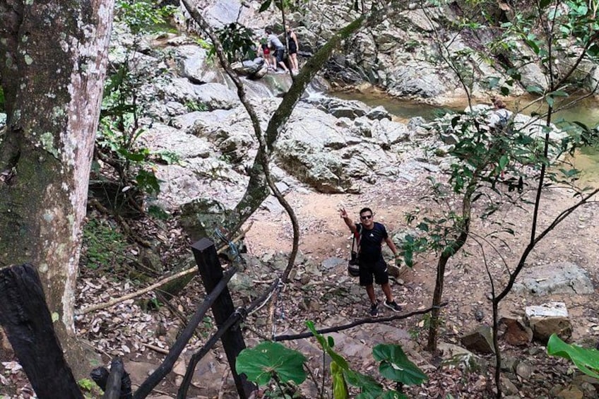 Une personne se tient sur un sentier rocheux, dans une zone boisée, avec des arbres et un ruisseau en arrière-plan. 4
