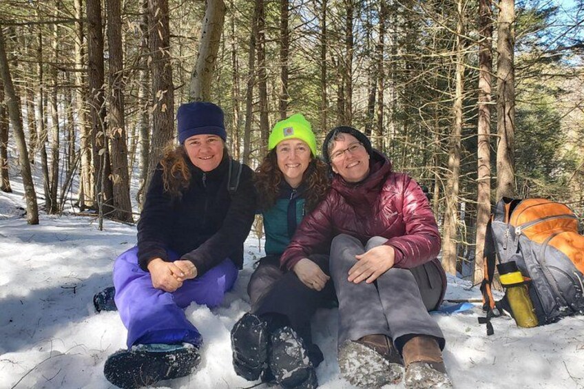 Three people sitting on the snow in a forest with trees and a backpack in the background. 4