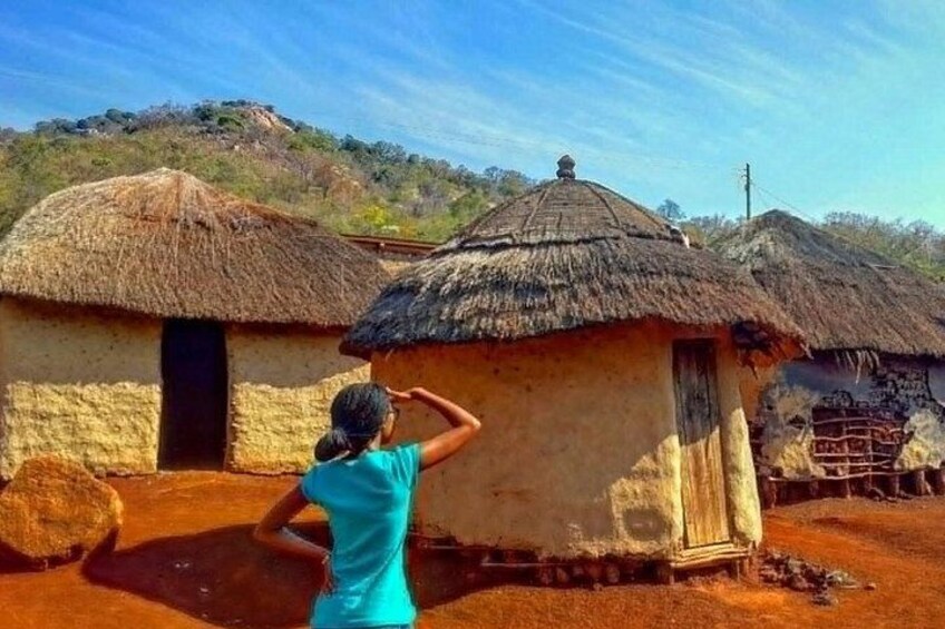 A person taking a photo of traditional thatched-roof huts with a hillside in the background. 4