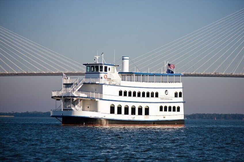 A white riverboat with multiple decks and a flag, sailing under a large suspension bridge. 2