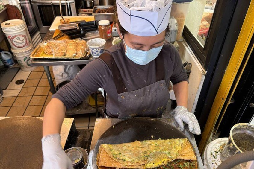 Una persona cocinando en una cocina, usando mascarilla y guantes, con una sartén grande y una bandeja de comida al fondo. 5