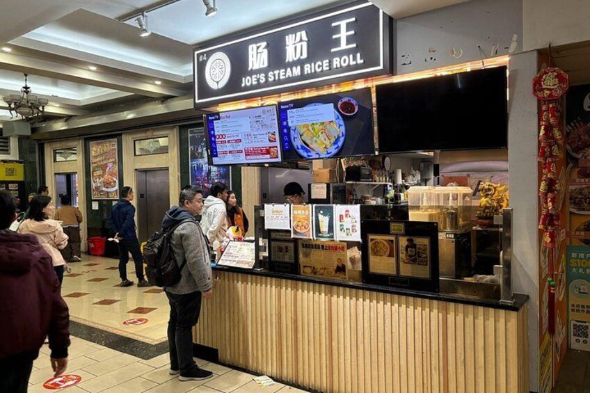 Un puesto de comida llamado 'Joe's Steamed Rice Roll' con un cartel de menú y una pantalla digital. 2