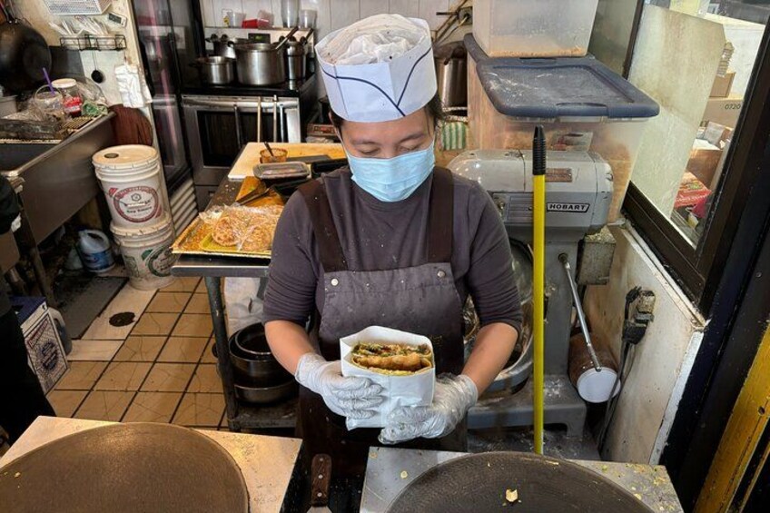 Una persona en una cocina preparando comida, usando mascarilla y guantes, con utensilios de cocina e ingredientes a la vista. 4