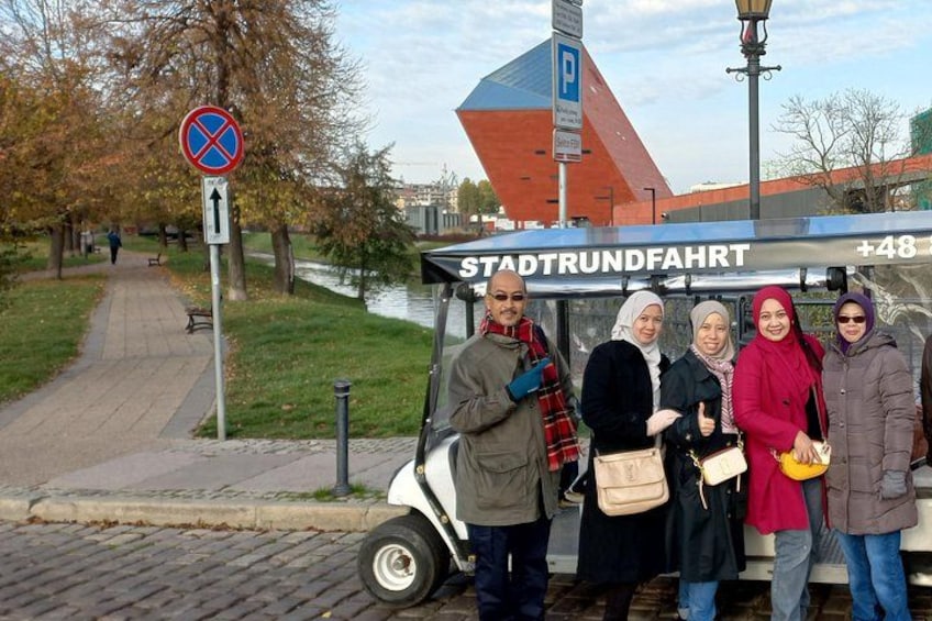 A tram with people standing beside it, a no parking sign, a pedestrian path, and a building with a red roof. 5