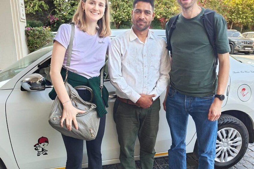 Drie personen staan voor een witte auto met een rode logo-sticker. 3
