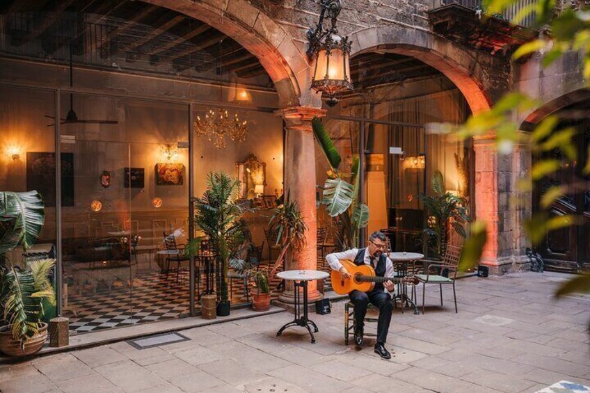Un músico tocando la guitarra en un café al aire libre con mesas y sillas, rodeado de plantas en macetas y bajo un amplio arco. 2
