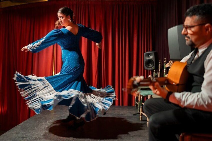 Una mujer con un vestido azul baila en el escenario mientras un hombre toca la guitarra al fondo. 3
