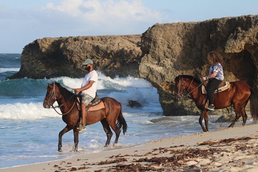 Deux personnes à cheval, galopant le long d’une plage où les vagues s’écrasent contre les rochers. 1