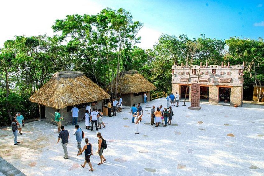A courtyard with thatched-roof huts, a stone building, and visitors. 1