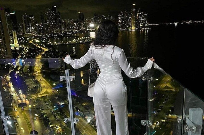 Une personne vêtue de blanc se tient sur un balcon, d’où elle a une vue sur le paysage urbain illuminé la nuit. 1