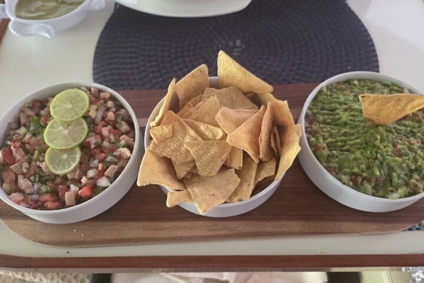 Una mesa de madera con tres recipientes de comida: un recipiente de salsa con cuñas de lima, un recipiente de totopos y un recipiente de guacamole con un totopo. 5
