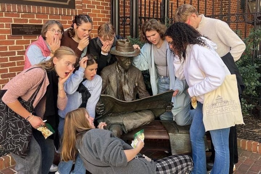 Un grupo de personas reunidas alrededor de una estatua, algunas sosteniendo bebidas, en un espacio al aire libre con paredes de ladrillo y una cerca metálica. 1