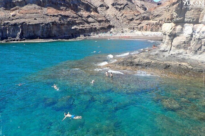 Una zona costera con aguas azules cristalinas, acantilados rocosos y gente nadando y tomando el sol. 3