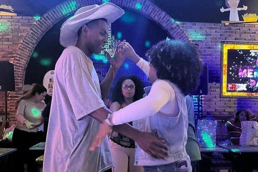 A person in a white hat and shirt dancing with another person in a dimly lit room with brick walls and neon signs. 2