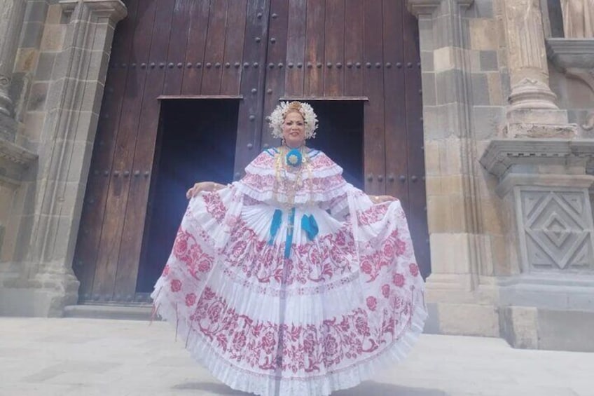 A person in a traditional white dress with pink embroidery, standing in front of a large wooden door with stone architecture. 1