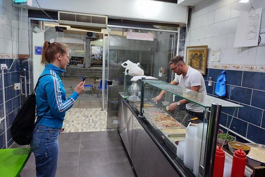 A person standing at a counter in a food establishment, another person behind the counter, and a menu board in the background. 4