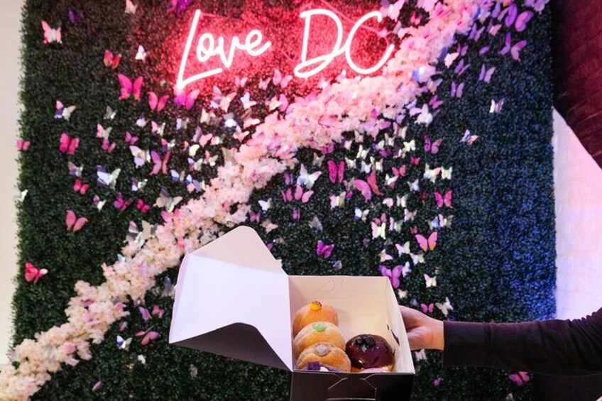 A person holding a box of donuts with a backdrop of flowers and a neon sign that reads "Love DC". 2