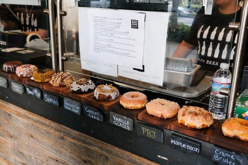 A variety of donuts displayed on a counter with labels such as 'Samosas', 'Chocolate Cup Cake', 'Vanilla Glazed', 'PBJ', and 'Maple Bacon'. 5