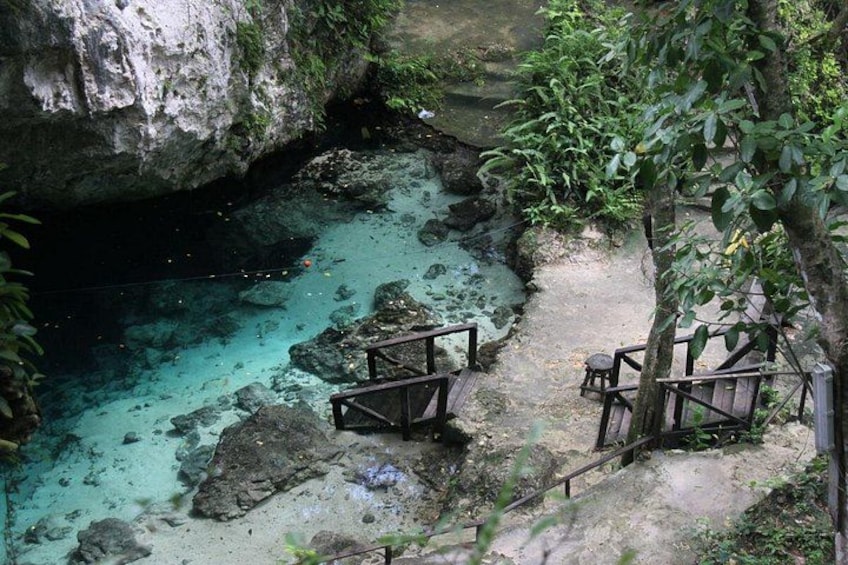 Entrada rocosa a una cueva con agua turquesa cristalina, escalones de madera y barandilla metálica. 2