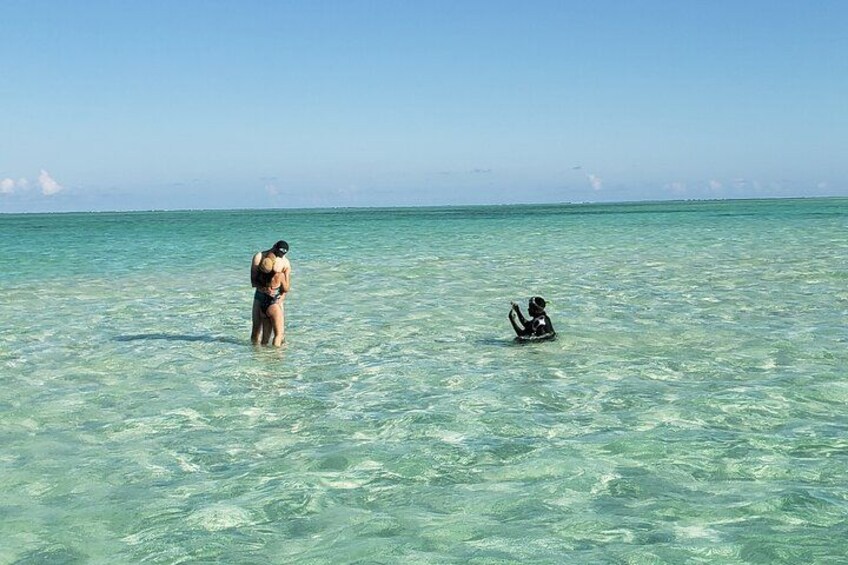 A person standing in shallow water, another person sitting on a surfboard, and a distant horizon. 4