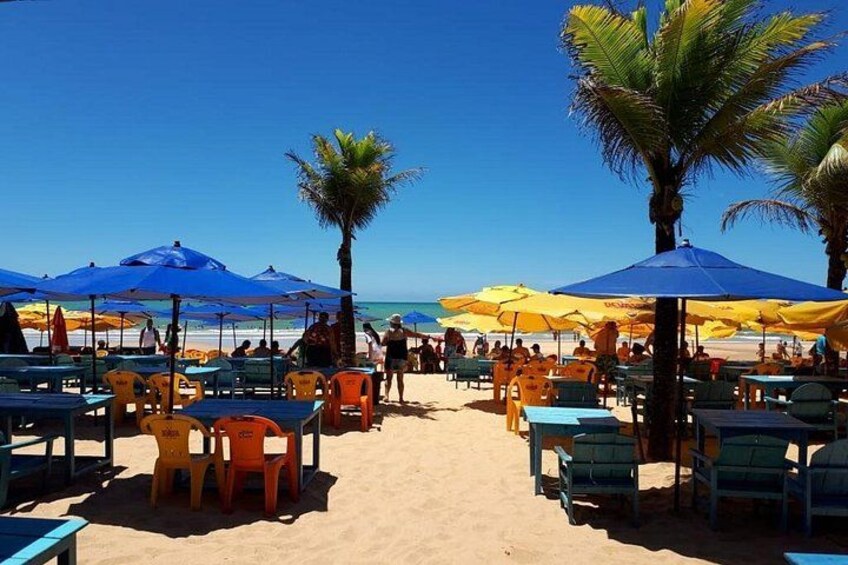 Ein Strand mit Tischen und Stühlen unter blau-gelben Sonnenschirmen, Palmen und Menschen. 3