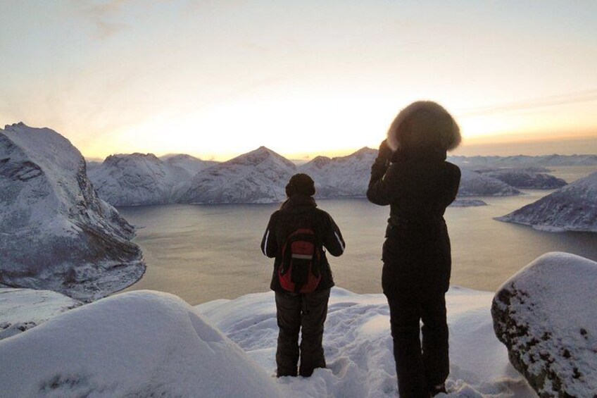 Deux personnes se tiennent sur une falaise enneigée, surplombant un plan d’eau et des montagnes, au moment du coucher du soleil. 2