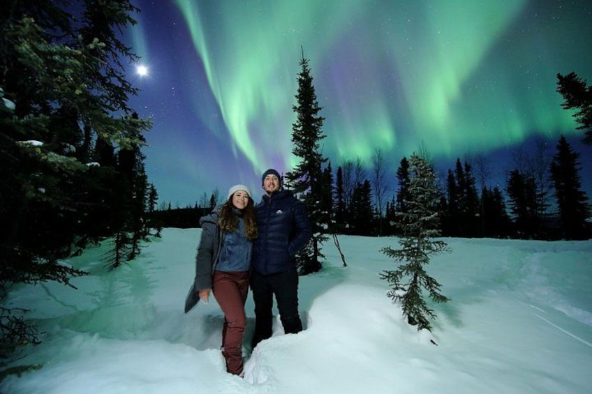 A couple standing in a snowy forest with the aurora borealis in the background. 1