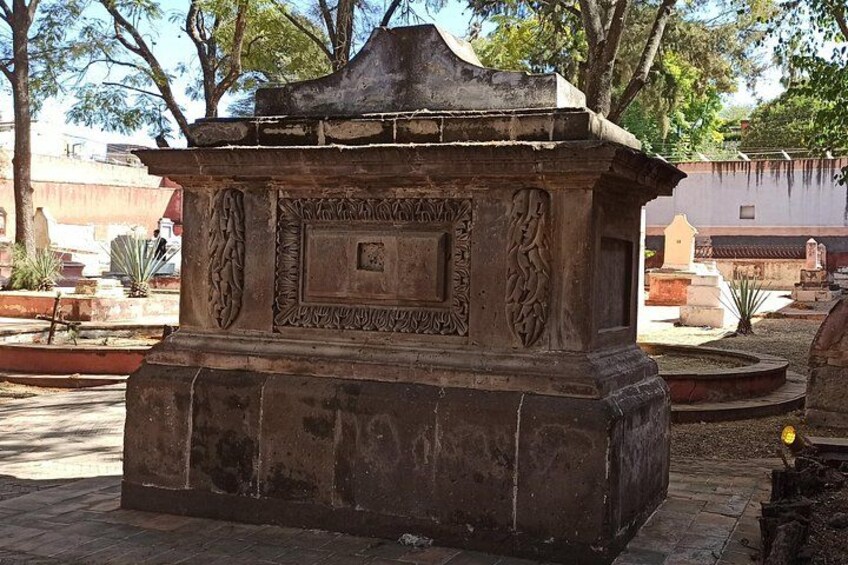 Un monumento de piedra con intrincados grabados y una placa, rodeado de árboles y otras lápidas en un cementerio. 3