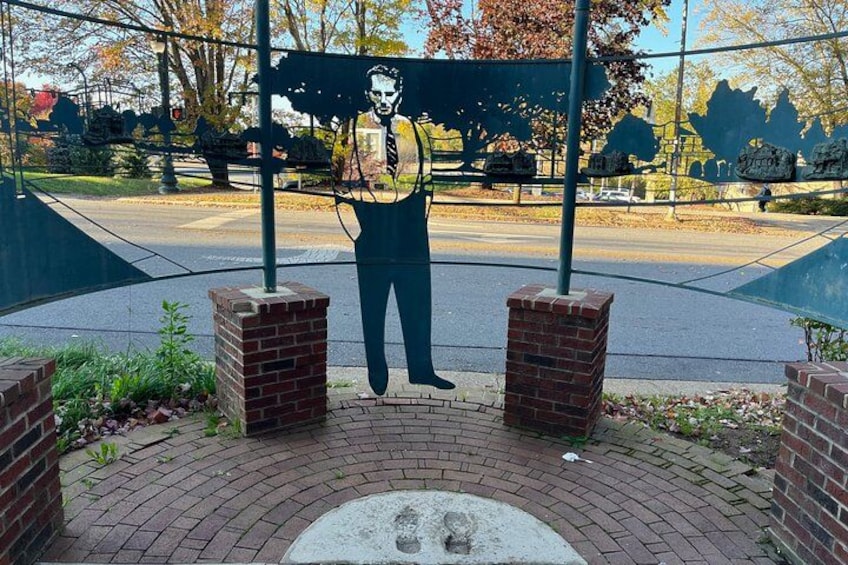 Una escultura de un hombre sosteniendo un cartel, rodeada de muros de ladrillo y un área pavimentada con árboles al fondo. 2