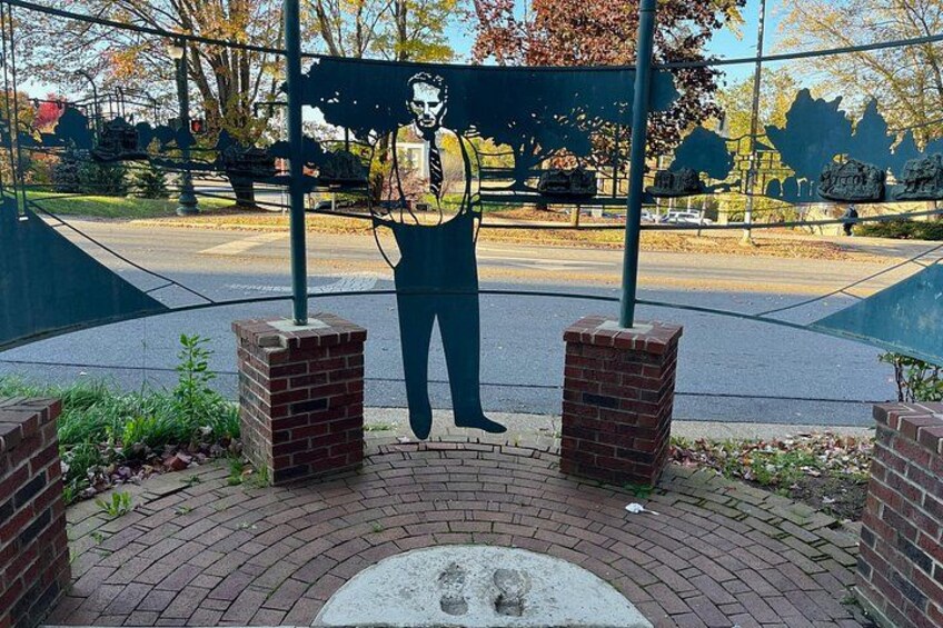Escultura de un hombre sosteniendo un libro, rodeado de muros de ladrillo y una zona pavimentada con árboles al fondo. 3