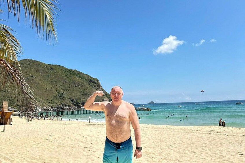 En strand med en person som spänner sina muskler, palmer och en brygga i bakgrunden. 2