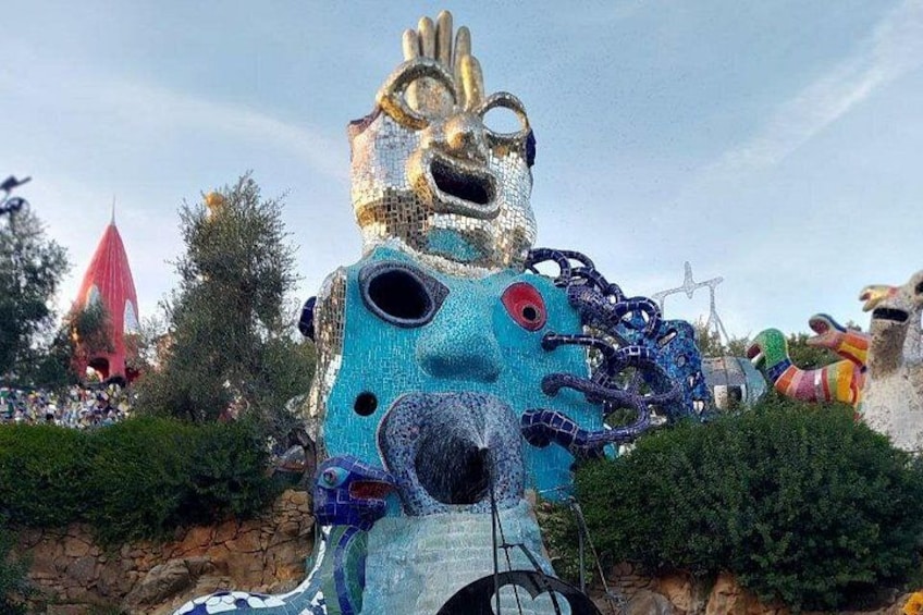A large, colorful mosaic sculpture of a face with multiple eyes and tentacles, set against a backdrop of trees and a red structure. 4