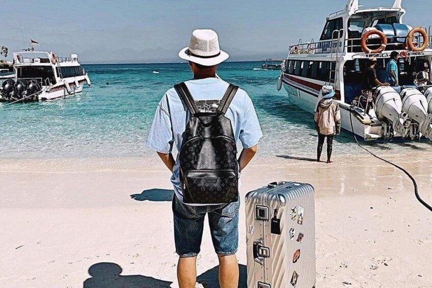 A man with a backpack and a suitcase on a beach, a boat in the water, and another boat docked with passengers. 5