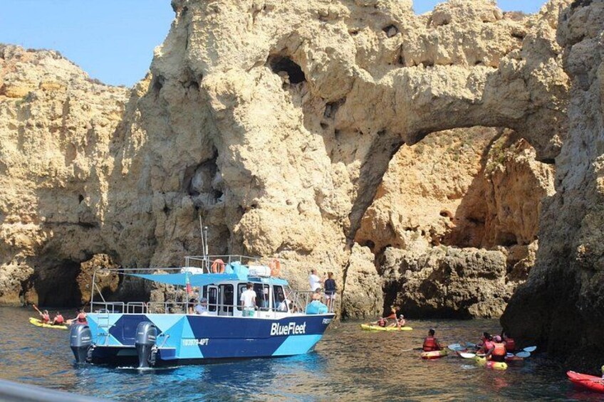 Una embarcación llamada 'BlueFish' con personas haciendo kayak en el agua y una formación rocosa en forma de arco al fondo. 2