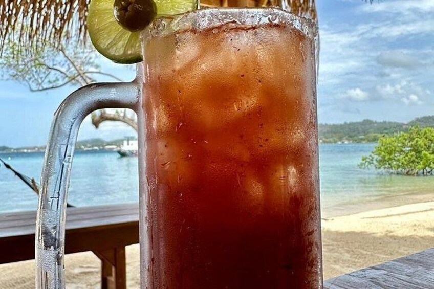 A tall glass with a dark liquid, ice cubes, and a lime wedge, set against a beach backdrop with clear skies. 2