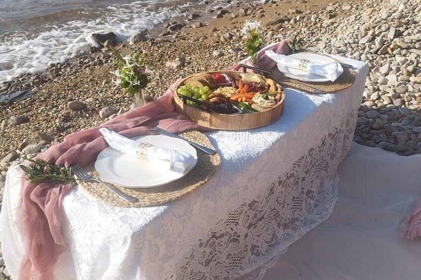 A picnic setup on a rocky beach with a table covered in lace, plates, and a basket of food. 2