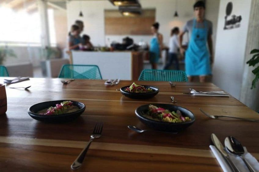Una mesa de madera con platos de comida, cubiertos y una persona con delantal azul al fondo. 4
