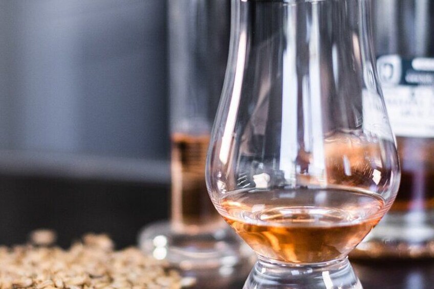 A glass of whiskey with a clear liquid and a golden hue, grains scattered on the table, and a blurred background with a bottle. 2
