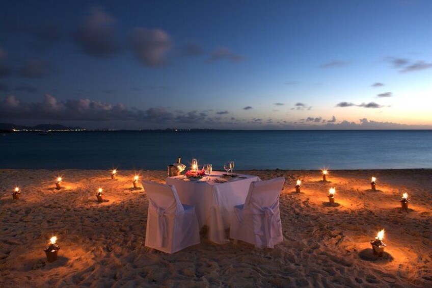 A beach dinner setup with a table and chairs, surrounded by candles. 2