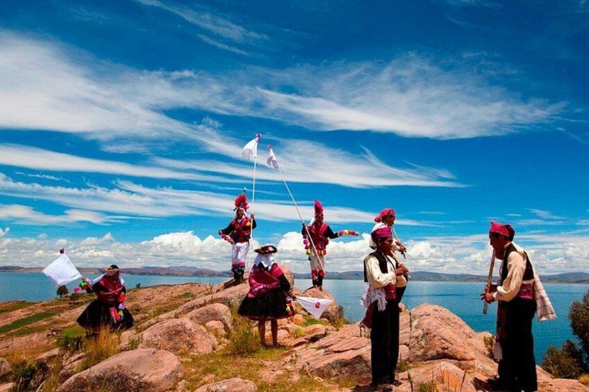 Un grupo de personas con vestimenta tradicional en una ladera rocosa, con cielo despejado y un cuerpo de agua al fondo. 2