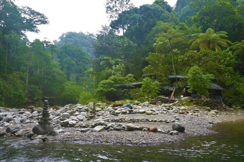 A riverbank with a stack of stones, lush greenery, and a small structure in the background. 1