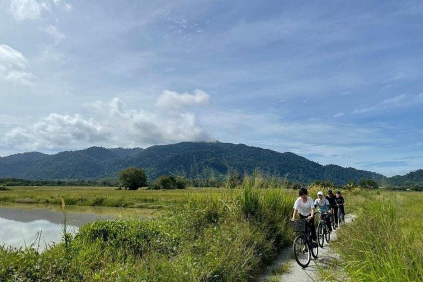 A group of cyclists on a narrow path beside a water body, with hills in the background. 3