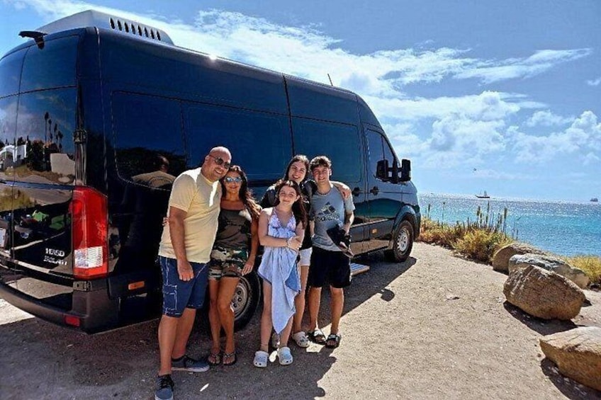 A group of people posing in front of a black van by a coastal road with large rocks and the sea in the background. 1